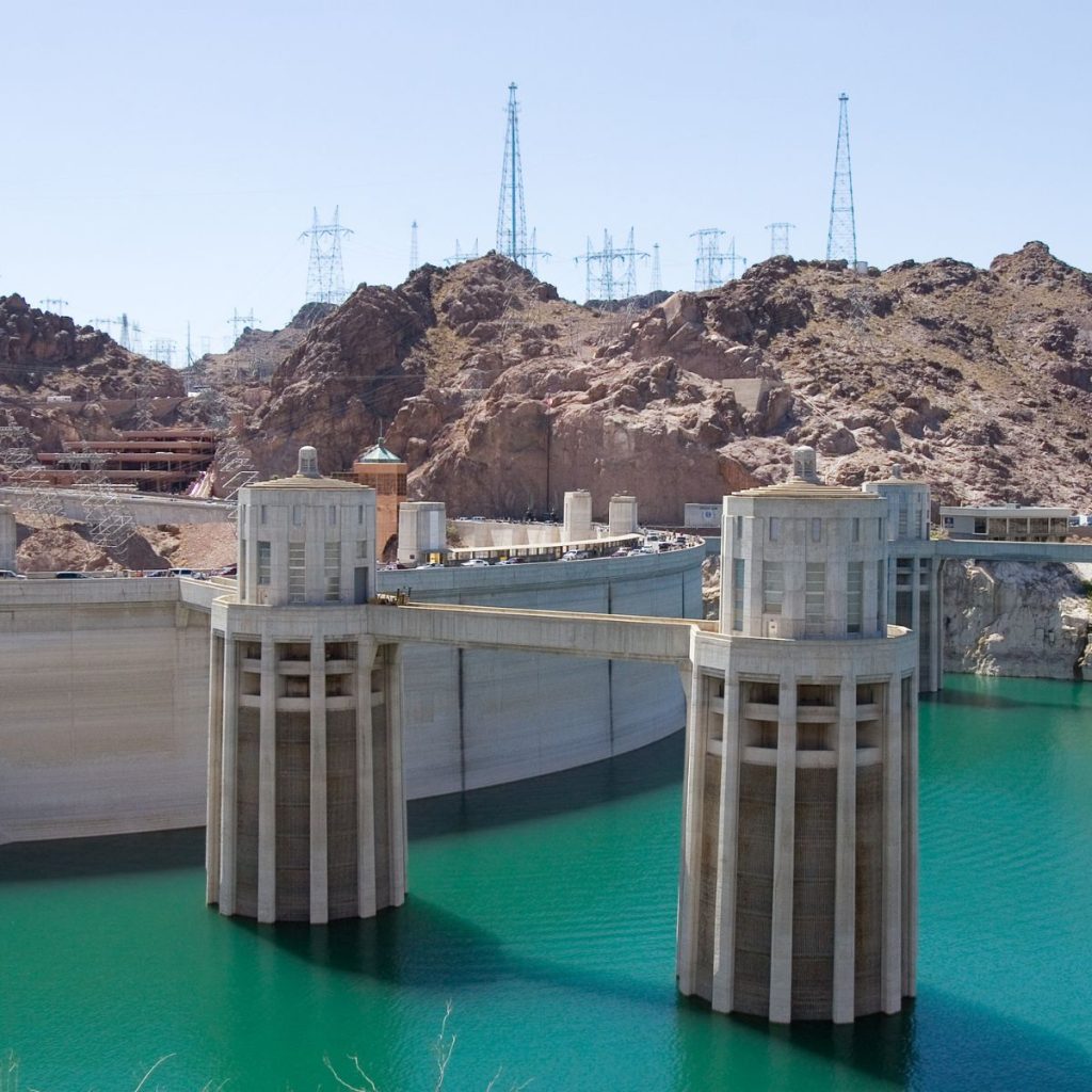 Hoover Dam complex at the beginning of Lake Mead from the Arizona side with a bridge over the Colorado River in the background and the surrounding rocky hills with power towers.