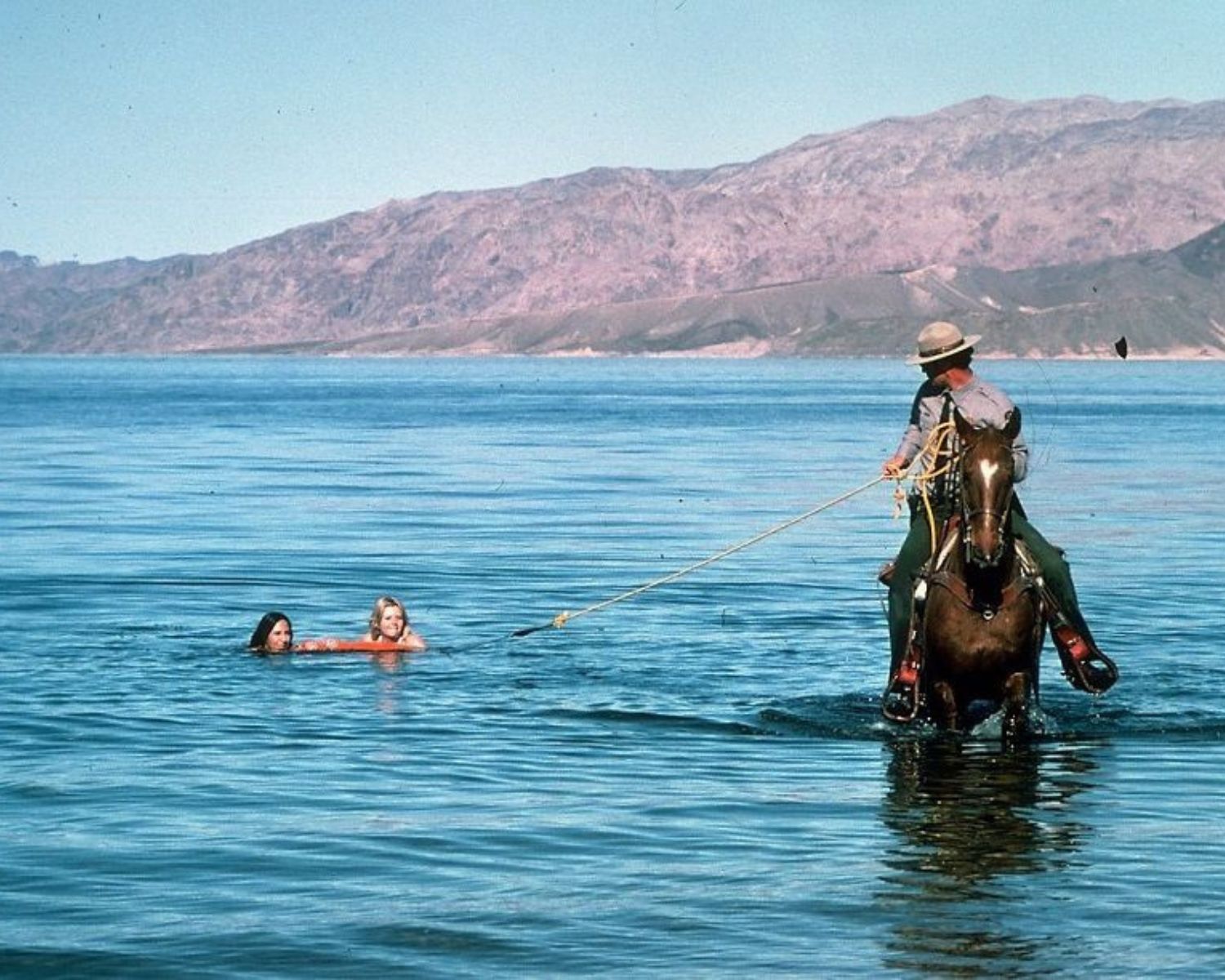 A Park Ranger on horseback lassos a personal watercraft used by two young girls in a desert lake.