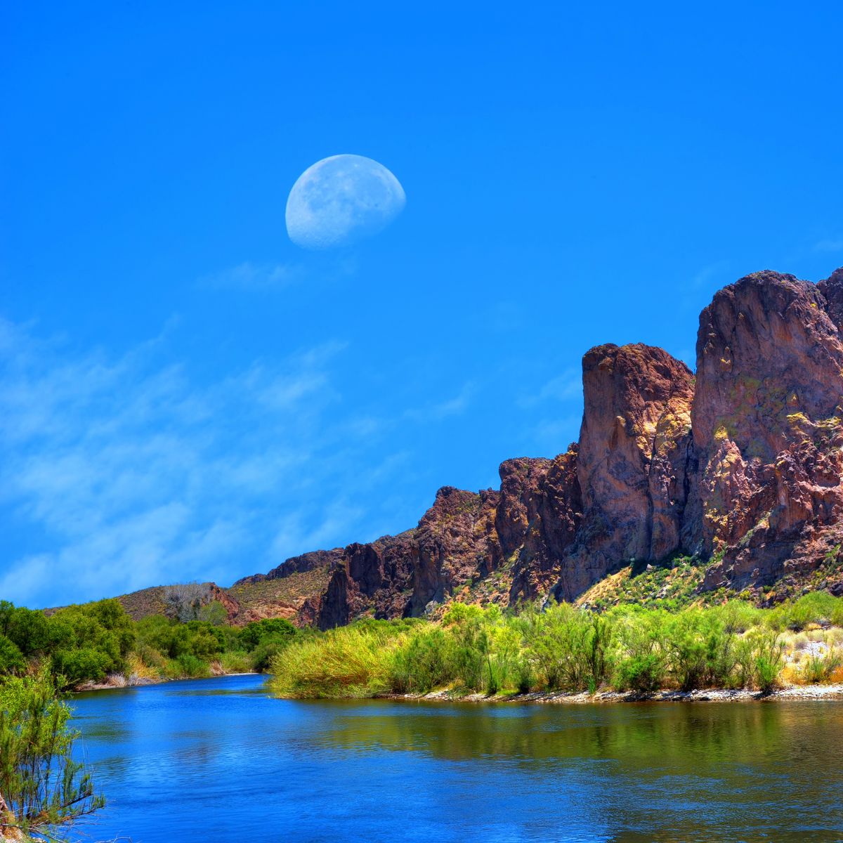 a peaceful river scene with the moon in the sky and rocky hills in the background.